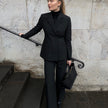 En kvinne kledd i en svart Avira-blazer for damer, en turtleneck og bukser står på en steintrapp og holder en svart håndveske.
