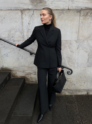 En kvinne kledd i en svart Avira-blazer for damer, en turtleneck og bukser står på en steintrapp og holder en svart håndveske.