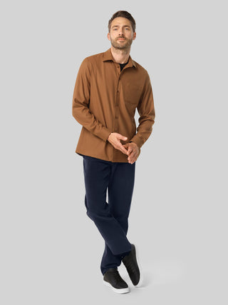 En mann kledd i en uformell skjorte i kamelfarge, mørke bukser og svarte joggesko.