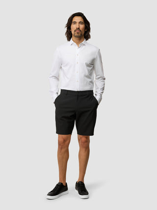 En mann kledd i en hvit skjorte med knapper, «Essential Shorts Black» og svarte joggesko.