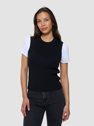 En kvinne som har på seg en svart, finstrikket ribbestrikket damevest over en hvit T-skjorte og mørke jeans.