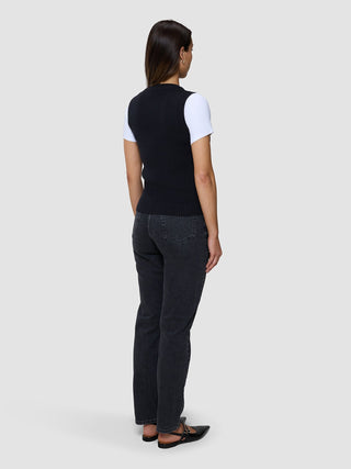 En kvinne sett bakfra, kledd i en svart, finstrikket ribbvest for kvinner over en hvit T-skjorte, mørke jeans og svarte ballerinasko.
