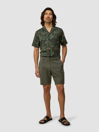 En mann kledd i en skjorte med botanisk mønster, GEN2™-shorts i dempet grønnfarge med 360°-stretch og svarte sandaler.