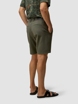 En person i GEN2™-shorts i dempet grønnfarge og en mønstret grønn skjorte, iført svarte sandaler i skandinavisk design.