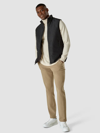 En smilende mann som har på seg en svart «Utility Vest» over en kremfarget turtleneck, med beige bukser og hvite joggesko.