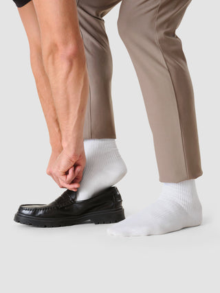 En person i beige bukser og hvite sokker (2-pakning) som tar på seg en svart loafer.