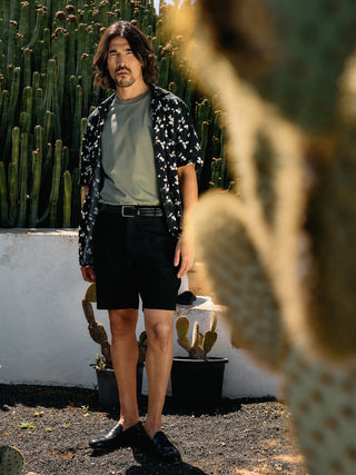 En mann i en grønn T-skjorte, en mønstret skjorte med knapper og svarte Essential Shorts står foran noen kaktuser.
