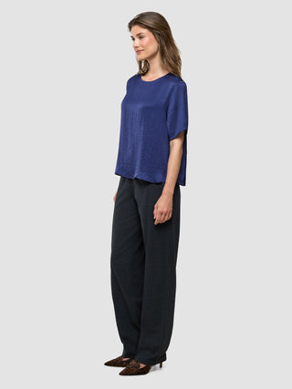 En kvinne viser frem «Amara Blouse Navy» for damer med krøllete tekstur, kombinert med mørke bukser og høyhælte sko med leopardmønster.