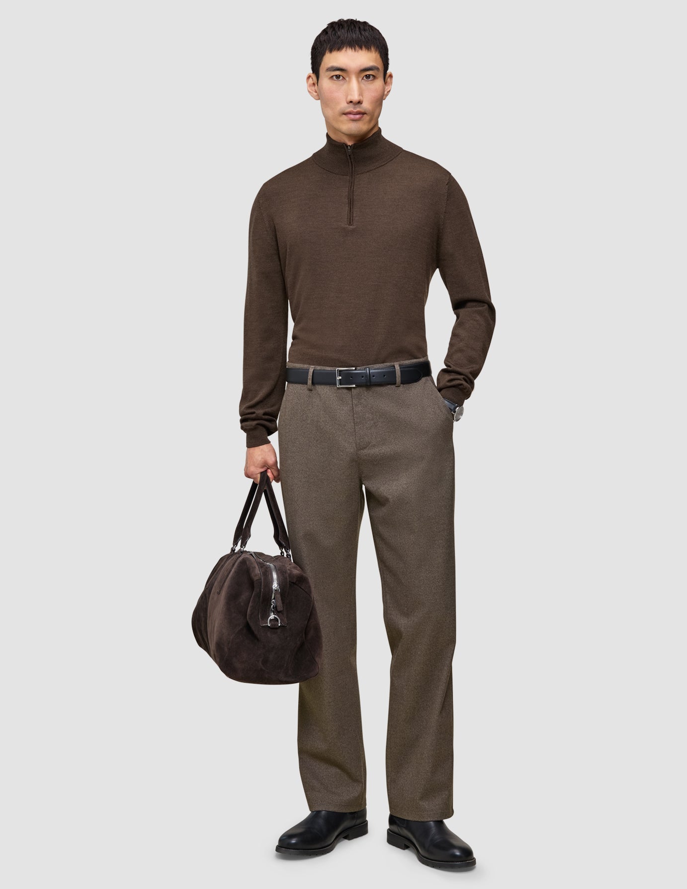 En mann i en brun genser med kvart glidelås og Atelier Pants Relaxed Dark Beige-bukser holder en mørk veske som fremhever skandinavisk design.