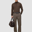 En mann i en brun genser med kvart glidelås og Atelier Pants Relaxed Dark Beige-bukser holder en mørk veske som fremhever skandinavisk design.