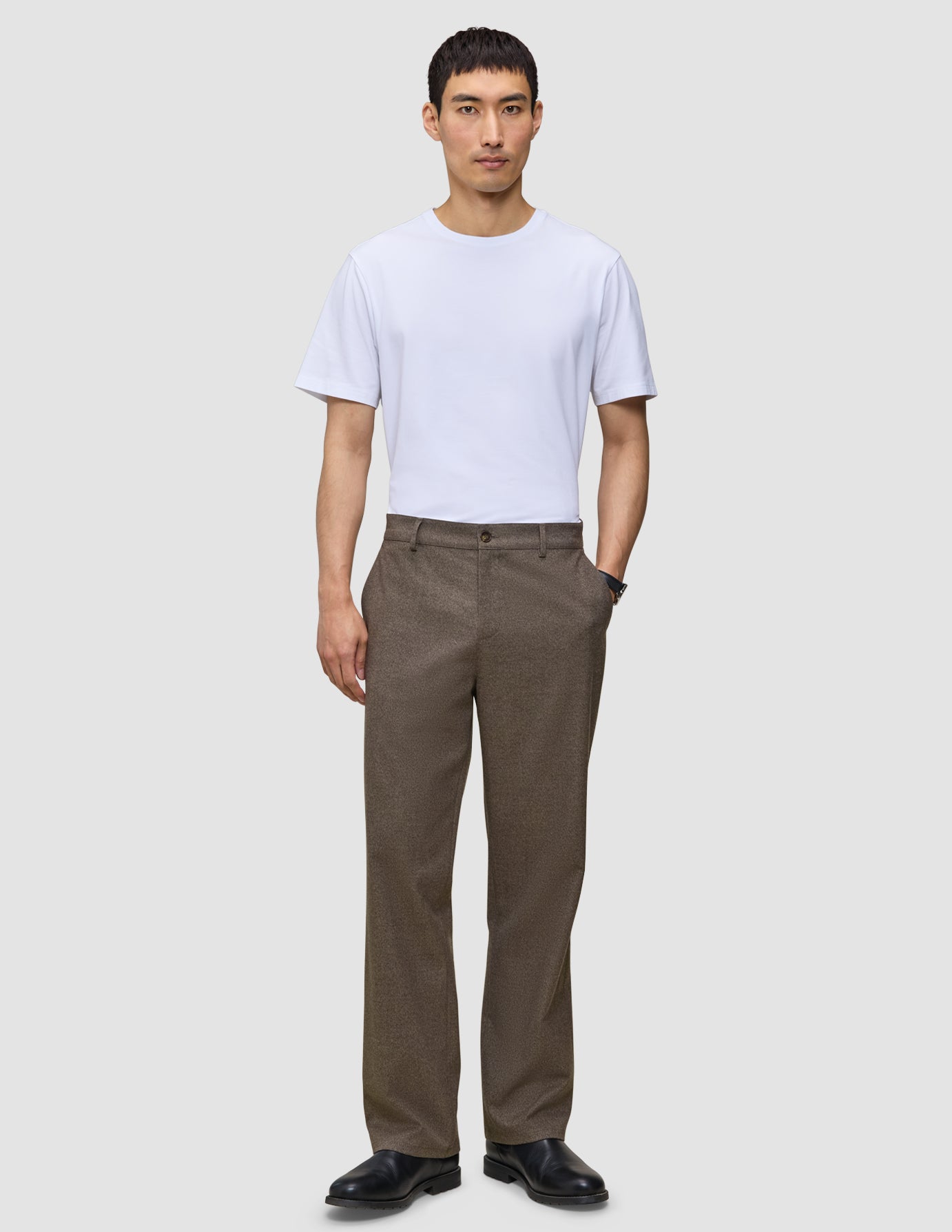 En mann har på seg en hvit T-skjorte, svarte sko og Atelier Pants Relaxed Dark Beige i brutt twill.