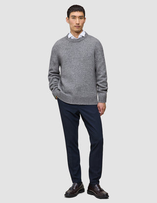 En mann kledd i en tykk, myk ullgenser med rund hals i fargen Glacier Grey over en hvit skjorte med krage, mørke bukser og brune loafers.