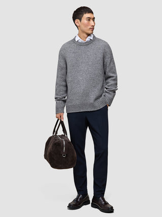 En mann kledd i en tykk, myk ullgenser med rund hals i fargen Glacier Grey over en skjorte med krage og bukser, som bærer en duffelbag.