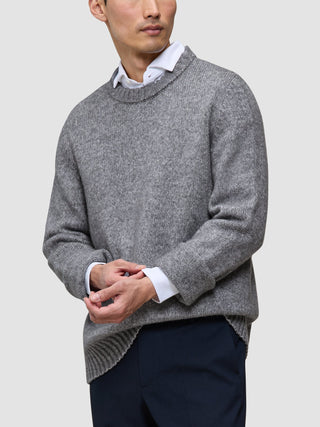 En person som har på seg en tykk, myk ullgenser med rund hals i fargen Glacier Grey over en hvit skjorte med krage og mørke bukser.