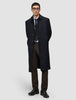 En mann i svart overfrakk, dressvest, skjorte og slips har på seg Motion Suit Pants Regular Cognac med hendene bak ryggen.