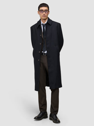 En mann i svart overfrakk, dressvest, skjorte og slips har på seg Motion Suit Pants Regular Cognac med hendene bak ryggen.