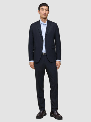 En mann kledd i Motion Suit-bukser i størrelse Regular i fargen Midnight Blue, med en lyseblå skjorte og mørke skinnloafers.