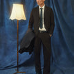 En mann kledd i Motion Suit Pants Relaxed Cognac, en hvit skjorte og et slips, som står ved siden av en gulvlampe.