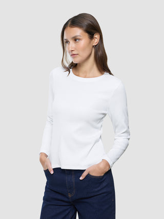 En kvinne iført en hvit «Women Rib»-T-skjorte med lange ermer og blå jeans, med hendene i lommene.