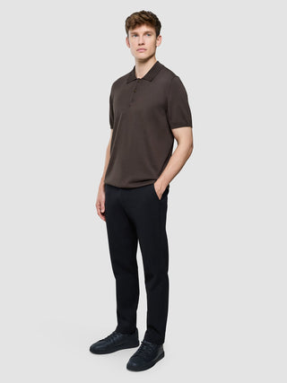 En mann kledd i en mørkebrun Seamless Core Knit-poloskjorte med korte ermer, svarte bukser og mørke joggesko.