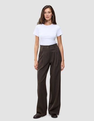 En kvinne har på seg en hvit T-skjorte og Serene Pants Wide Walnut Herringbone, som har et minimalistisk mørkebrunt design med smale striper.