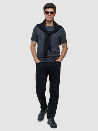En mann med solbriller, en «Supima Legacy»-T-skjorte i fargen «Deep Grey» med en genser over skuldrene og svarte 360°-stretchjeans.
