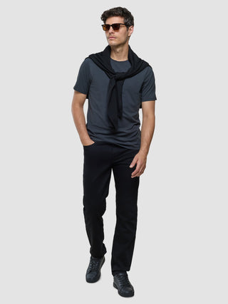 En mann i en Supima Legacy-T-skjorte i fargen «Deep Grey», svarte jeans, joggesko og solbriller, med en genser over skuldrene.