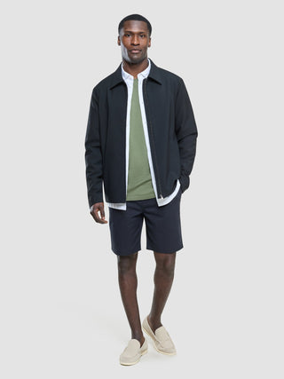 En mann i en svart jakke med glidelås over en hvit skjorte og en Supima-T-skjorte i fargen Clover Green, med mørke shorts og loafers.