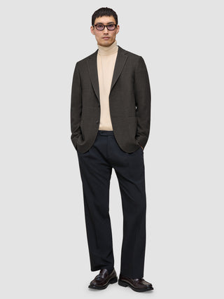 En person som har på seg en Timeless Wool Blazer Slim i mørkebrun rutemønster over en beige turtleneck og mørke bukser.