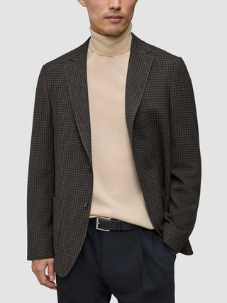 En person har på seg en Timeless Wool Blazer Slim i mørkebrun rutemønster over en beige turtleneck og svarte bukser.