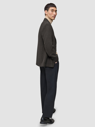 En mann kledd i en Timeless Wool Blazer Slim i mørkebrun rutemønster over en beige turtleneck og mørke bukser, som ser seg tilbake.