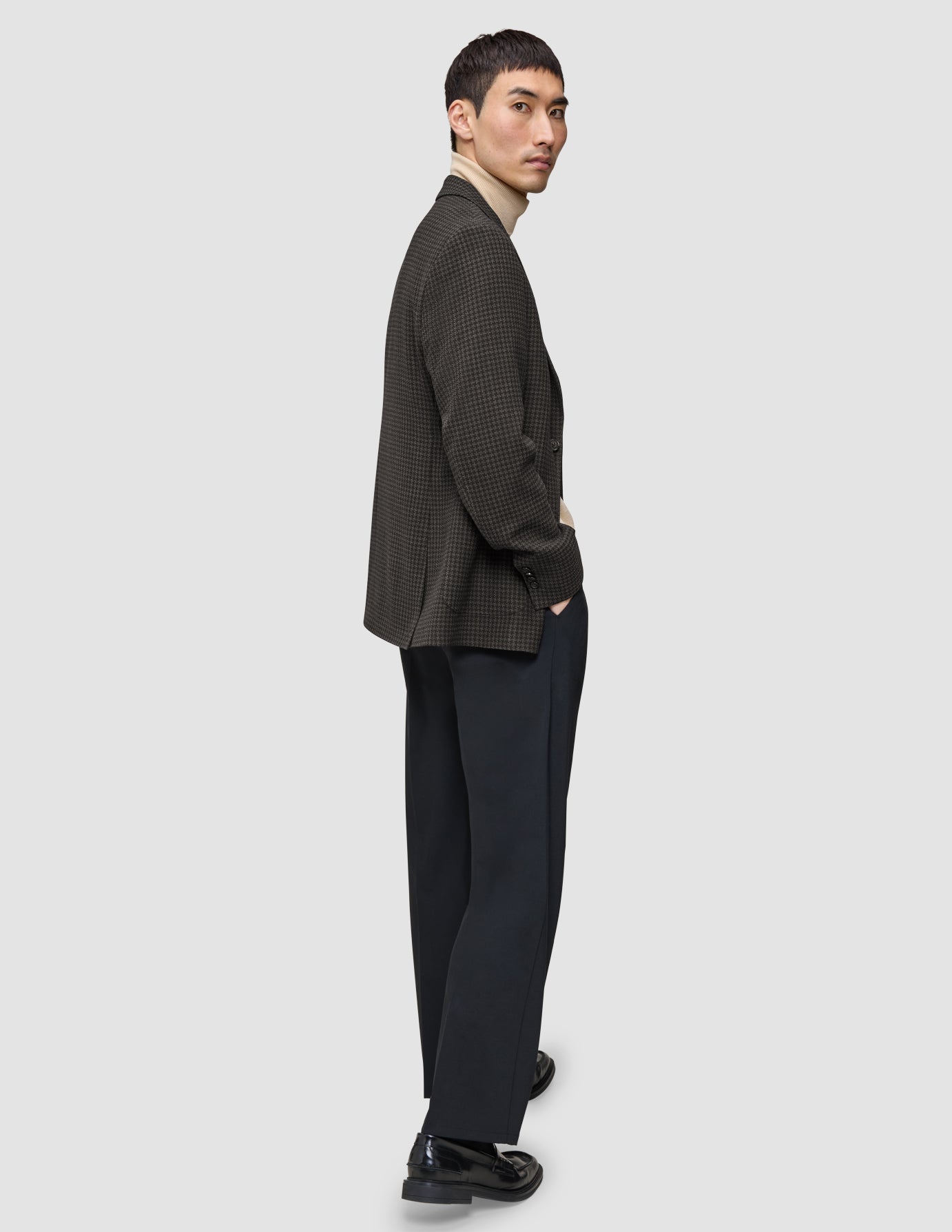 En mann kledd i en Timeless Wool Blazer Slim i mørkebrun rutemønster over en beige turtleneck og mørke bukser, som ser seg tilbake.