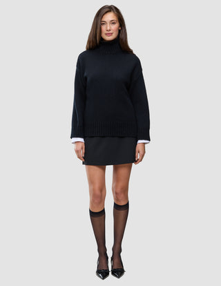En person som har på seg en svart, tykk og myk ullstrikket turtleneck for kvinner over en hvit skjorte, et svart skjørt og gjennomsiktige knestrømper.