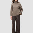 En person kledd i en tykk, myk ullstrikket turtleneck for kvinner i fargen taupe melange og brune bukser med smale striper.