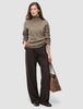 En kvinne kledd i en tykk, myk ullstrikket turtleneck for kvinner i fargen taupe melange og mørkebrune bukser med smale striper, som bærer en totebag.