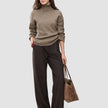 En kvinne kledd i en tykk, myk ullstrikket turtleneck for kvinner i fargen taupe melange og mørkebrune bukser med smale striper, som bærer en totebag.