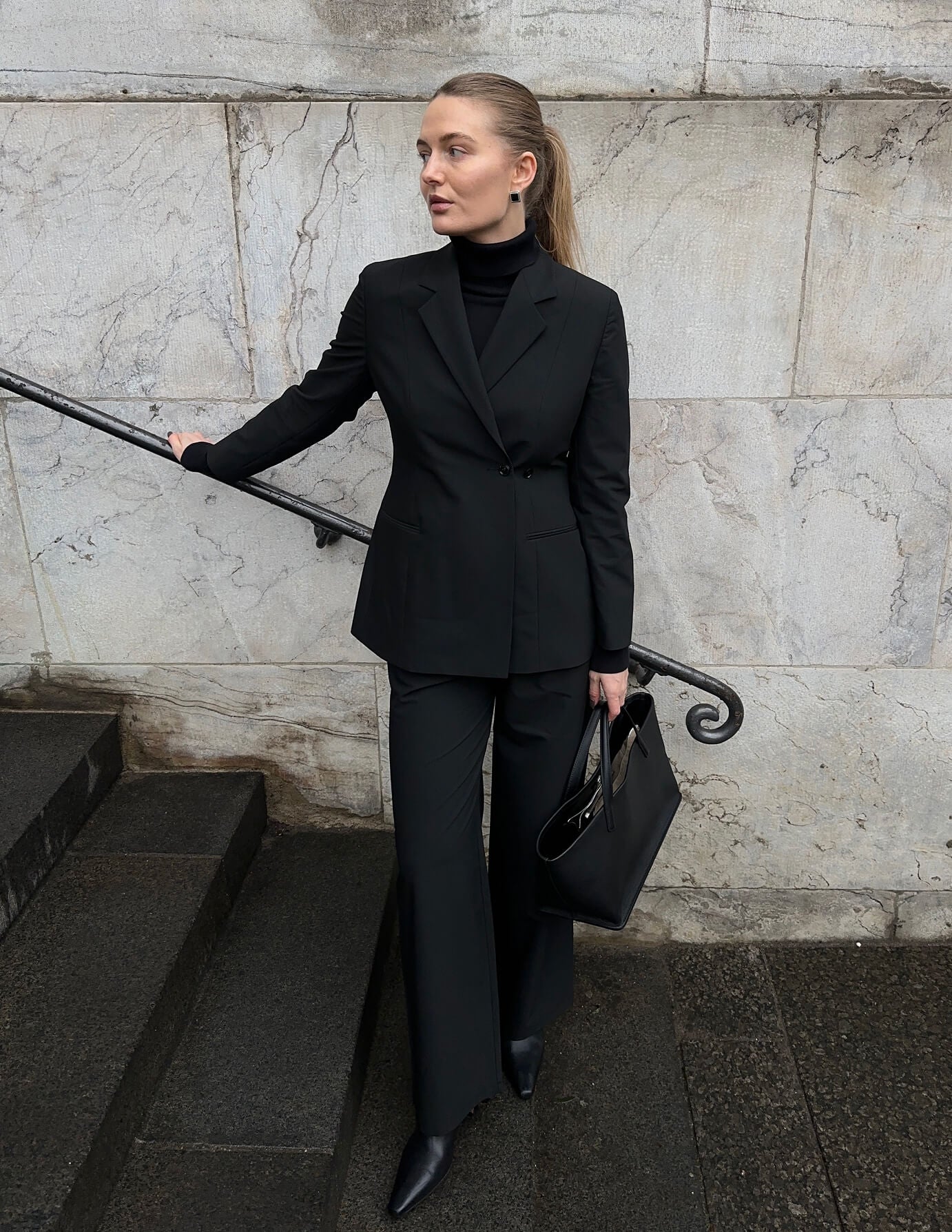 En kvinne kledd i en svart Avira-blazer for damer, en turtleneck og bukser står på en steintrapp og holder en svart håndveske.