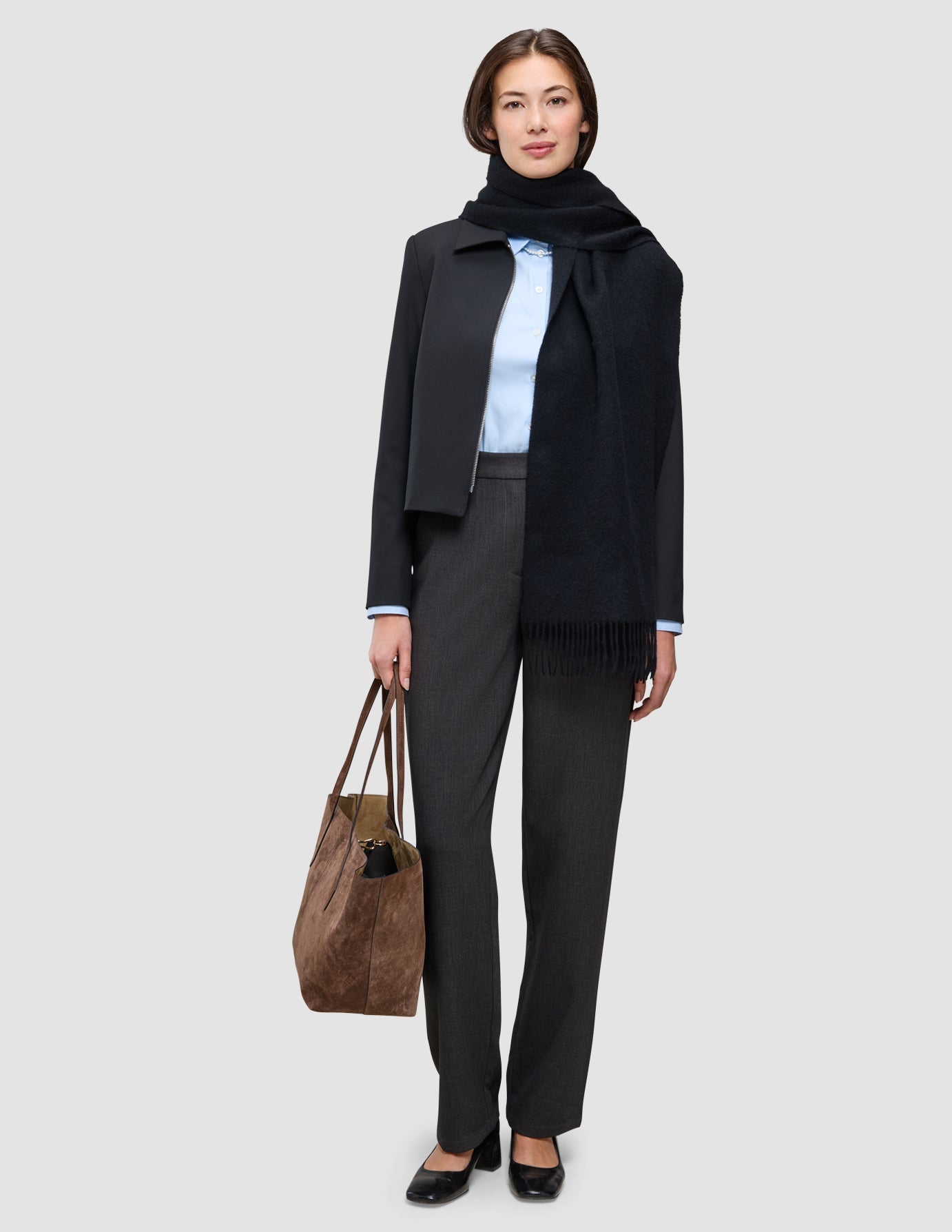 En kvinne som har på seg en Essential Zip Jacket fra An easy office upgrade over en Oxford-skjorte, grå bukser og en brun totebag.