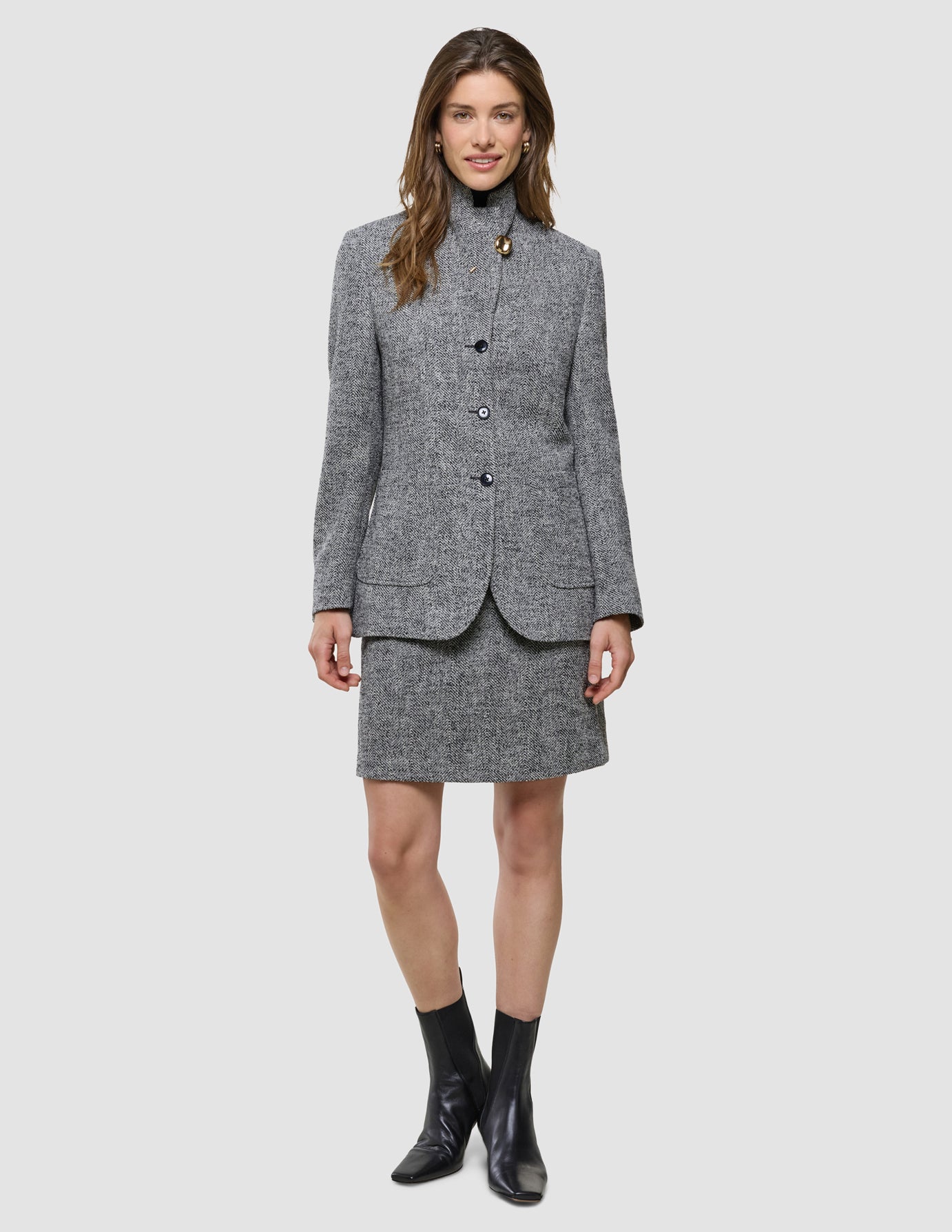 En kvinne kledd i en grå bouclé-blazer med fiskebensmønster, «Women Ashryn Short Skirt Domino Herringbone» og svarte ankelstøvler.