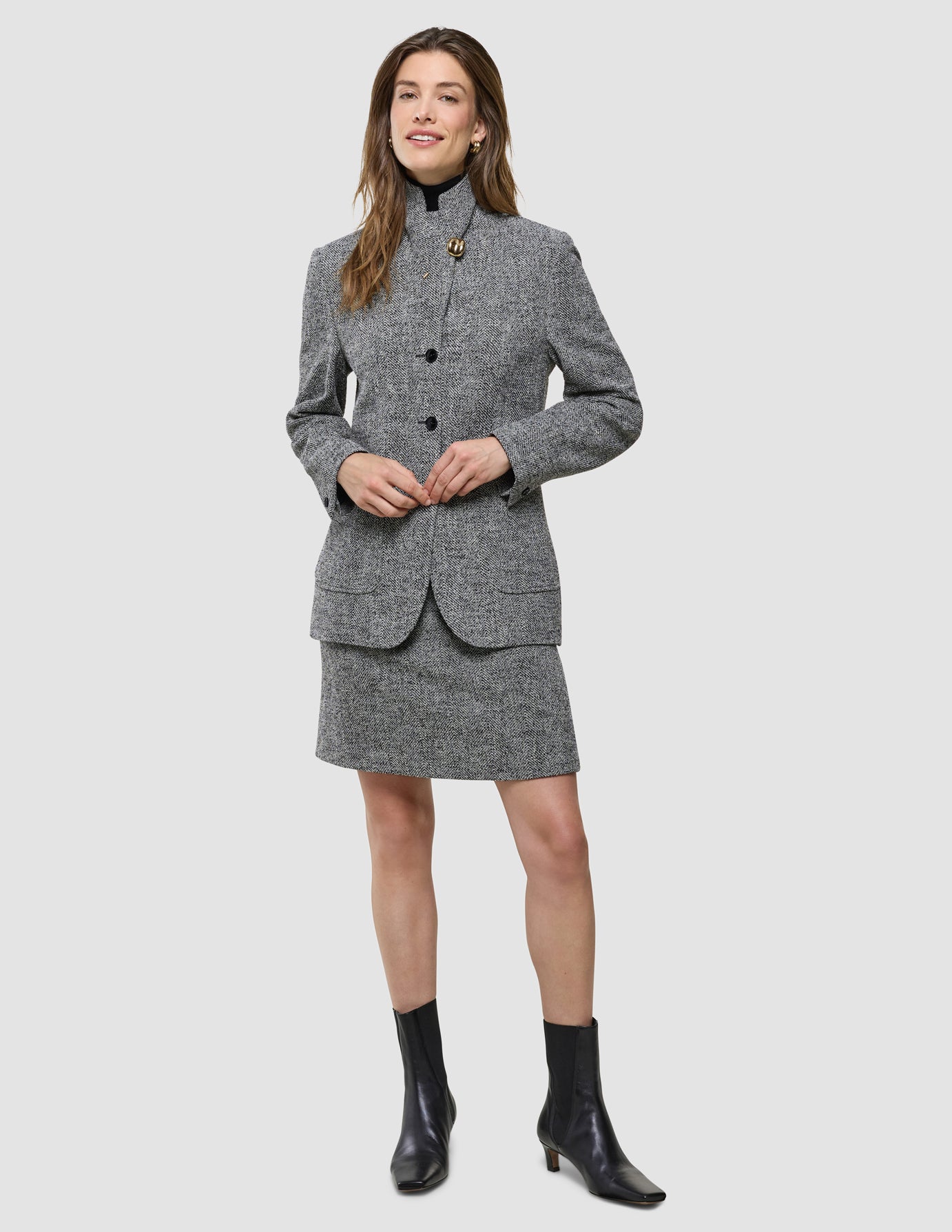 En kvinne kledd i en grå blazer med fiskebeinmønster, «Women Ashryn Short Skirt Domino Herringbone», og svarte ankelstøvler.