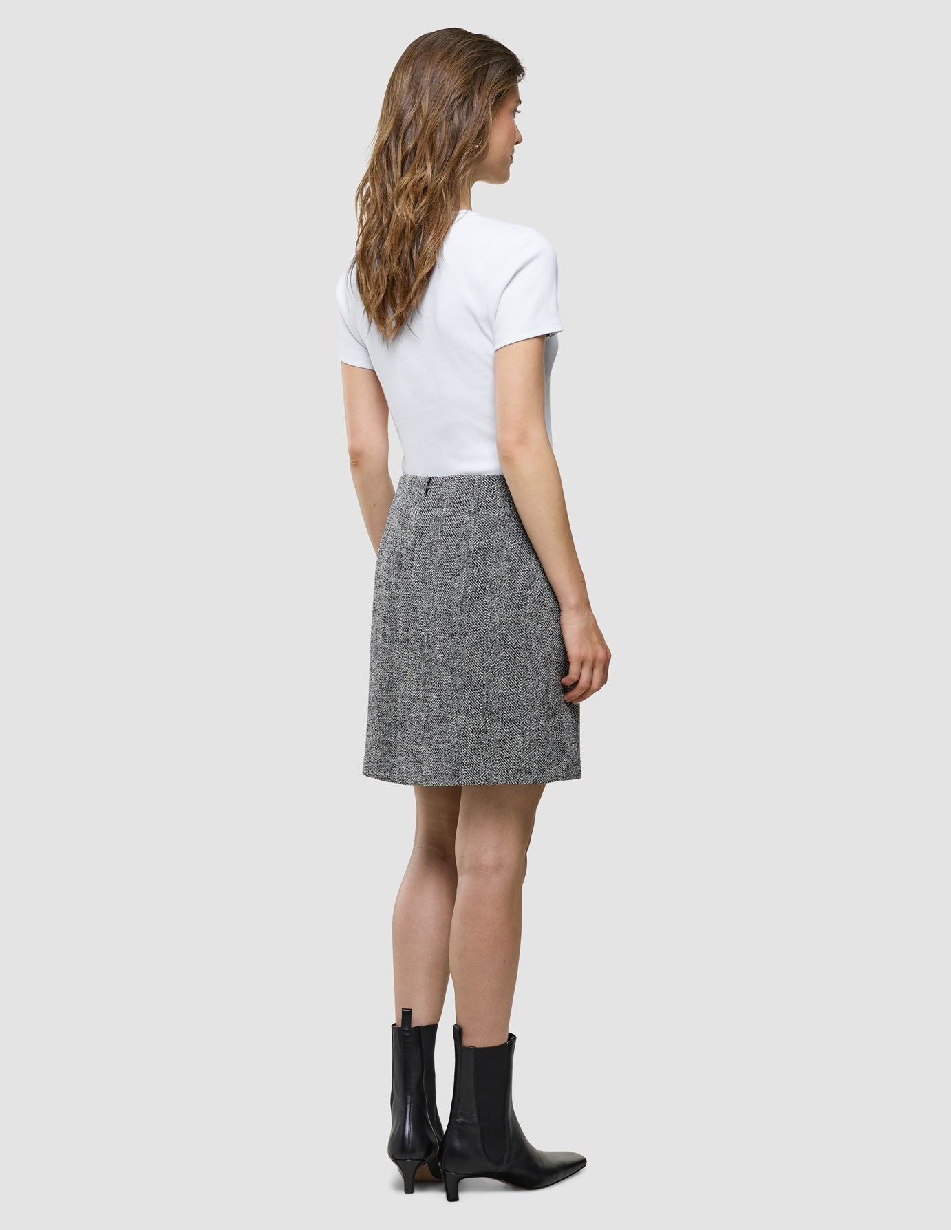 En kvinne sett bakfra, kledd i en hvit T-skjorte, et «Women Ashryn Short Skirt Domino Herringbone»-skjørt og svarte ankelstøvler.