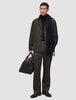 En mann i mørkebrune Atelier Pants Relaxed, svart genser og skjerf, som holder en svart skinnreiseveske.