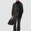 En mann i mørkebrune Atelier Pants Relaxed, svart genser og skjerf, som holder en svart skinnreiseveske.
