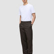 En mann i en hvit T-skjorte og mørkebrune Atelier Pants Relaxed, med hendene i lommene.