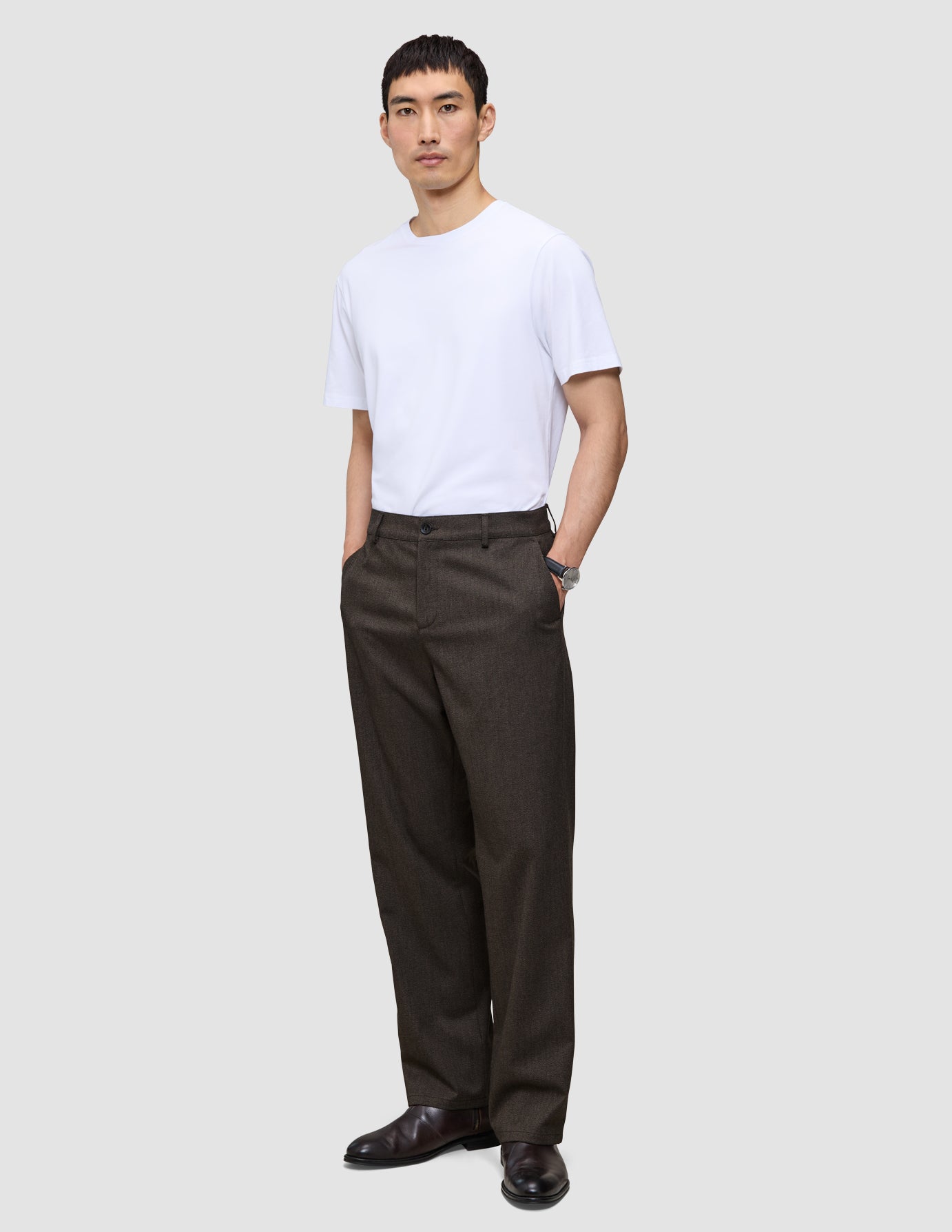 En mann i en hvit T-skjorte og mørkebrune Atelier Pants Relaxed, med hendene i lommene.