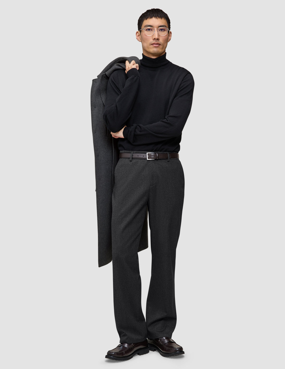 En mann i svart turtleneck og mørkegrå Atelier Pants Relaxed, som holder en frakk over skulderen på en avslappet måte.