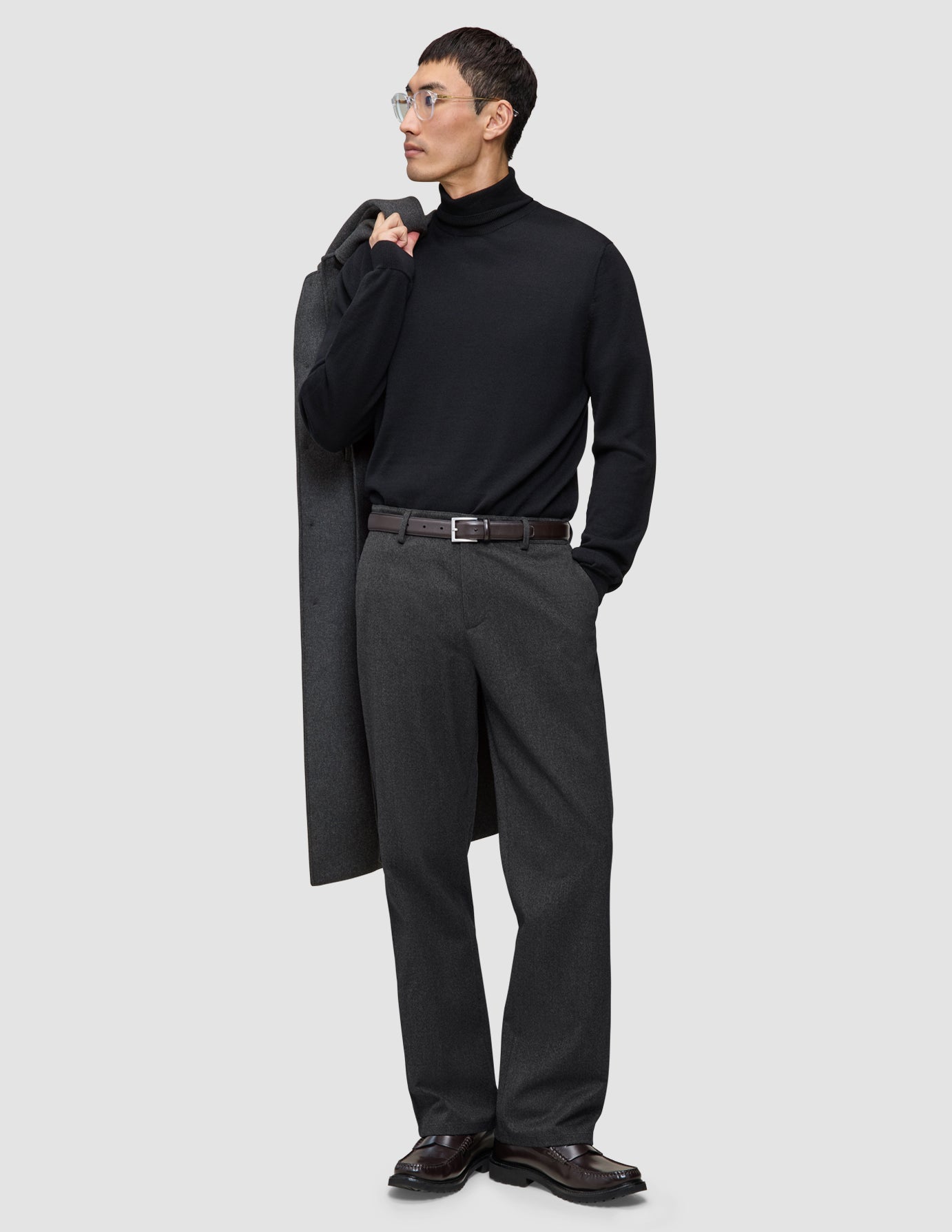 En mann i svart turtleneck og mørkegrå Atelier Pants Relaxed, med en jakke i skandinavisk design over skulderen.