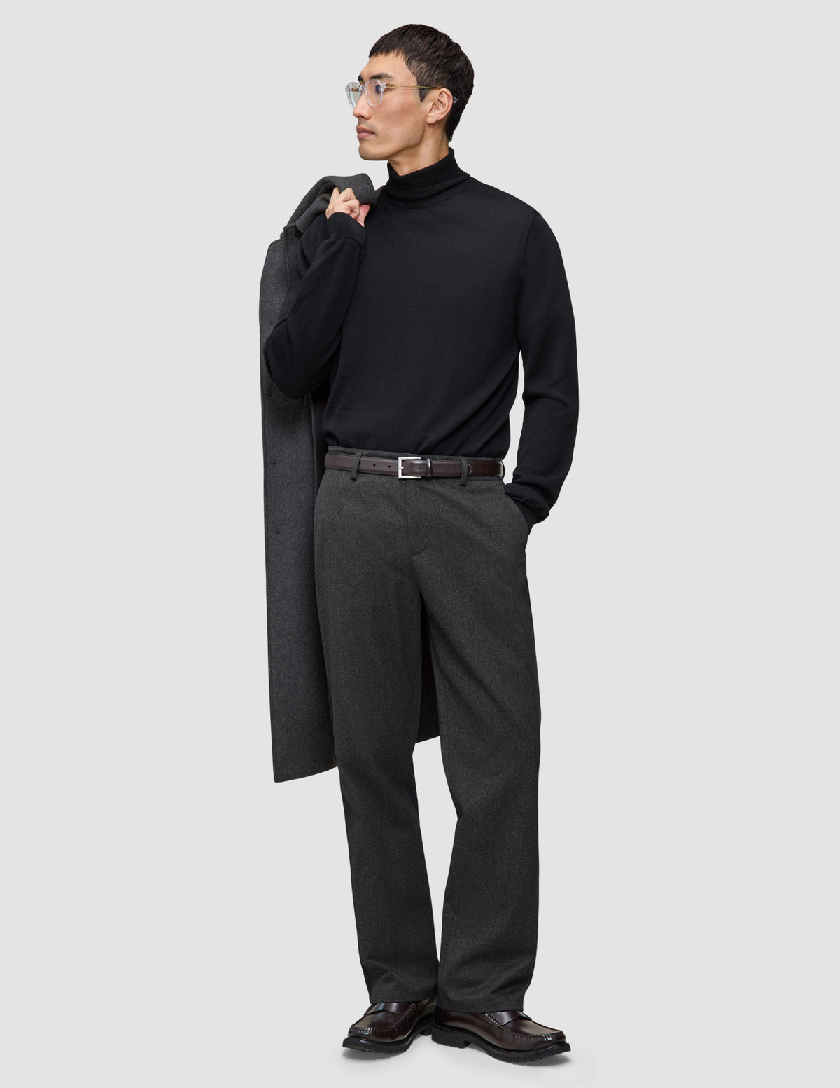 En mann i svart turtleneck og mørkegrå Atelier Pants Relaxed, med en jakke i skandinavisk design over skulderen.