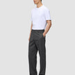 En person i en hvit T-skjorte og mørkegrå Atelier Pants Relaxed-bukser står med den ene hånden i lommen.