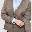 En tykk, myk ullstrikket damekardigan i taupe-melange, båret over en lyseblå skjorte med knapper.